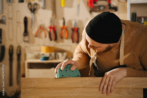 Attentive carpenter in a watch cap grinding a piece of wood, blurred background