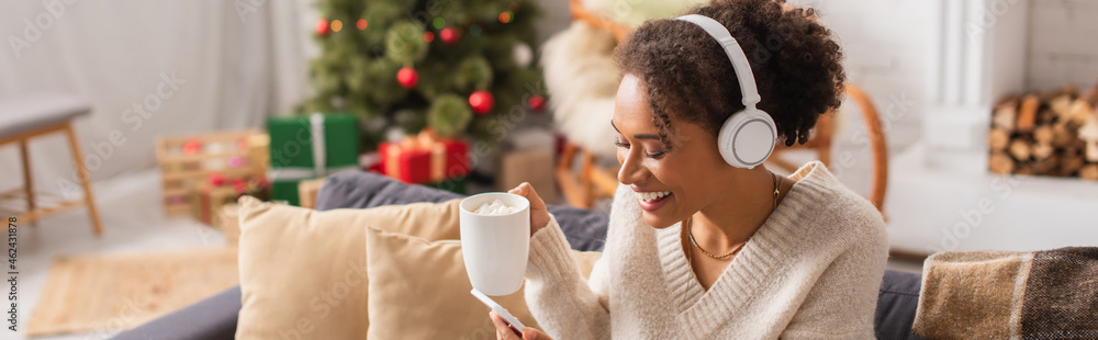 Cheerful african american woman in headphones using smartphone and holding cup during christmas at home, banner Cheerful african american woman in headphones using smartphone and holding cup during christmas at home, banner