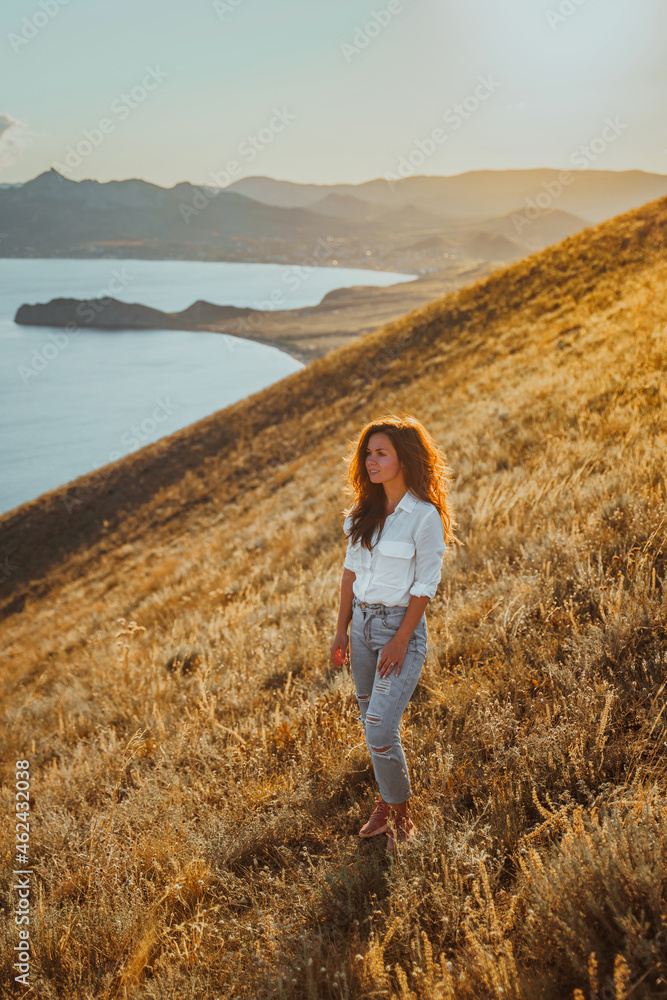 Beautiful young woman with long hair on top of a mountain at sunset with amazing views of the mountain slopes and the sea. The concept of an active lifestyle, freedom and achievement of goals.