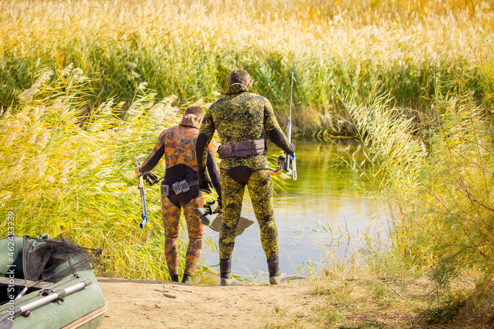 A fisherman in a diving suit stands on the bank of the river with a diving ammunition. Spearfishing, a man in a neoprene underwater suit, camouflaged clothing, a gun, diver camouflage with a harpoon. A fisherman in a diving suit stands on the bank of the river with a diving ammunition. Spearfishing, a man in a neoprene underwater suit, camouflaged clothing, a gun, diver camouflage with a harpoon.