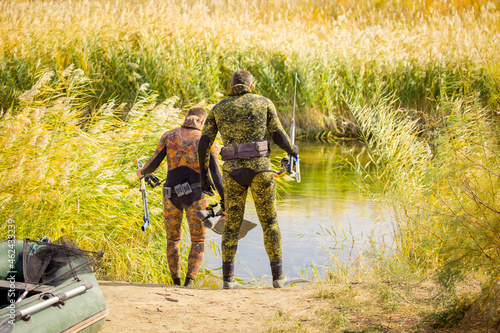A fisherman in a diving suit stands on the bank of the river with a diving ammunition. Spearfishing  a man in a neoprene underwater suit  camouflaged clothing  a gun  diver camouflage with a harpoon.