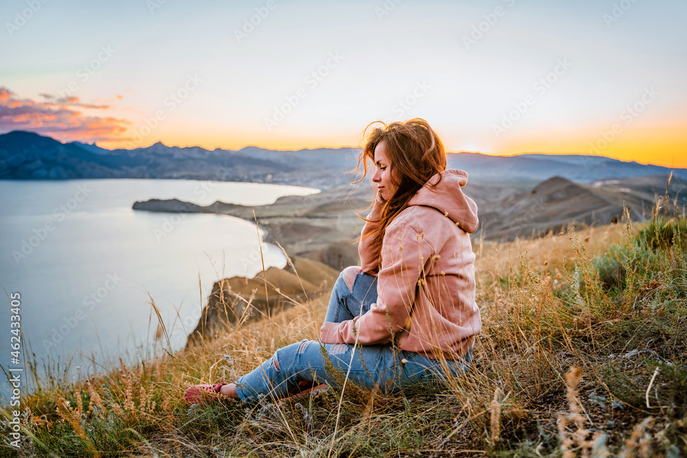 A beautiful young woman tourist is sitting on top of a mountain and enjoying the view of the sunset sea and mountains. The concept of freedom and meditation, an active lifestyle.