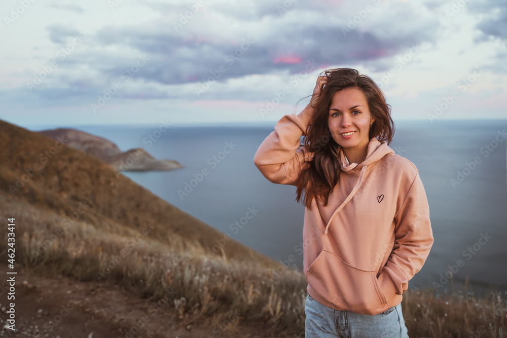 Beautiful young woman with long hair on top of a mountain at sunset with amazing views of the mountain slopes and the sea. The concept of an active lifestyle, freedom and achievement of goals.