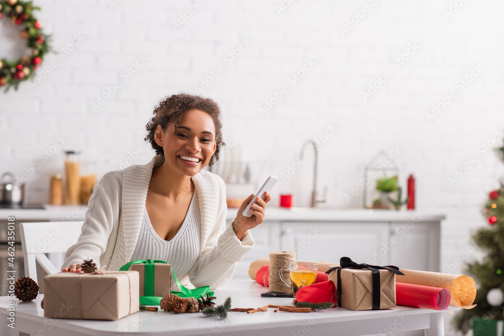 Positive african american woman holding cellphone near presents, christmas decor and tea Positive african american woman holding cellphone near presents, christmas decor and tea