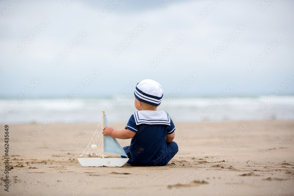 Baby boy sitting on the beach near the water and plays with a toy ship and bear toy Baby boy sitting on the beach near the water and plays with a toy ship and bear toy