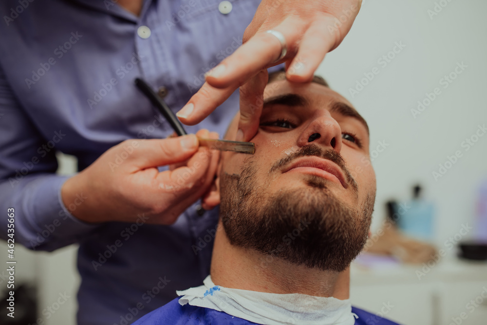 Fototapeta premium Close up portrait of handsome young man getting beard shaving with straight razor. Focus on the blade. Selective focus