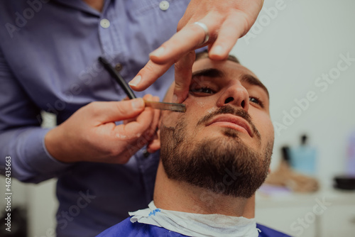Close up portrait of handsome young man getting beard shaving with straight razor. Focus on the blade. Selective focus