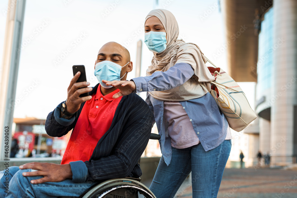 Muslim woman and disabled man wearing face mask using cellphone together Muslim woman and disabled man wearing face mask using cellphone together