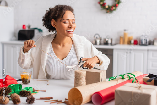 Positive african american woman cutting twine near decor and gifts at home