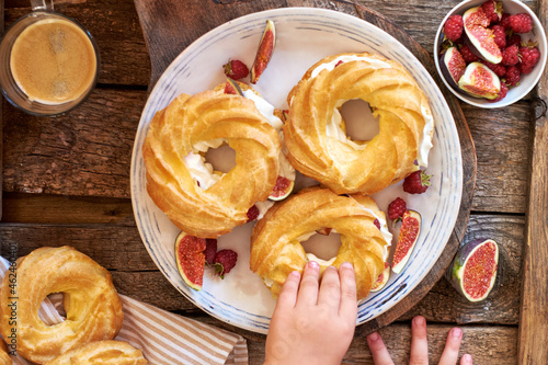 Choux rings with cream, figs and raspberries. Wooden background, top view. Children's hand holding a choux ring. © Lyudmila