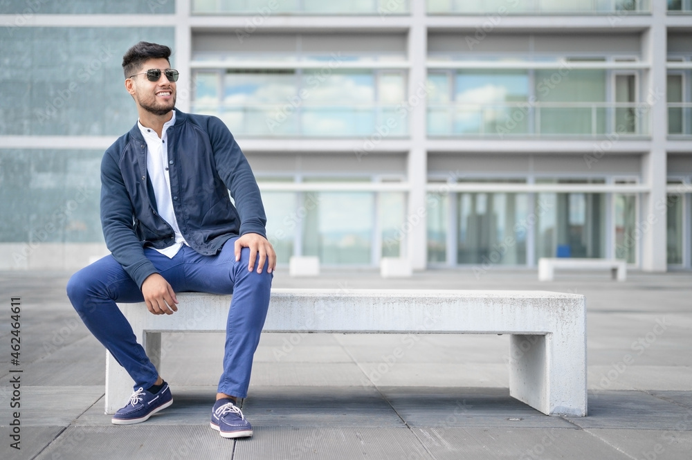Portrait of young man in stylish sunglasses posing while sitting in bench in urban background. High quality photo Portrait of young man in stylish sunglasses posing while sitting in bench in urban background. High quality photo
