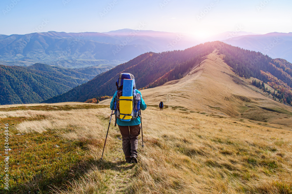 A girl in colored clothes with a large backpack walks along a beautiful mountain range in the Carpathians during a sunset