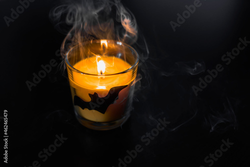 Halloween mystical decor with a lit candle in a glass beaker depicting a bat on a black background. Halloween holiday mystical smoke from a candle.
