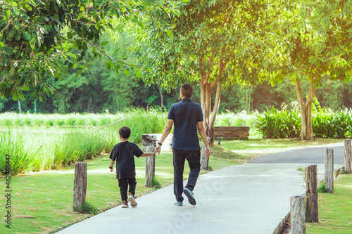 Asian father hand holding lovely son walking on pathway through green garden.Dad and son walking together in park. Happy family spending time together outside in green nature.Enjoying nature outdoors.