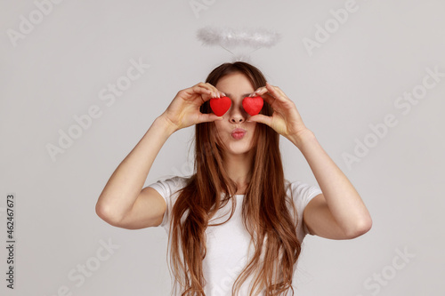 Woman covers eyes with toy hearts as if looking with love, sending air kiss, affection in relations, wearing white casual style T-shirt. Indoor studio shot isolated on gray background.