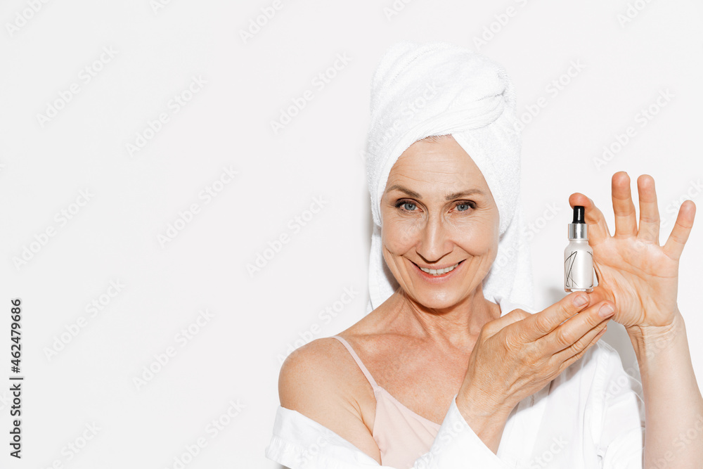 Mature woman wrapped in shower towel smiling while showing face serum Mature woman wrapped in shower towel smiling while showing face serum