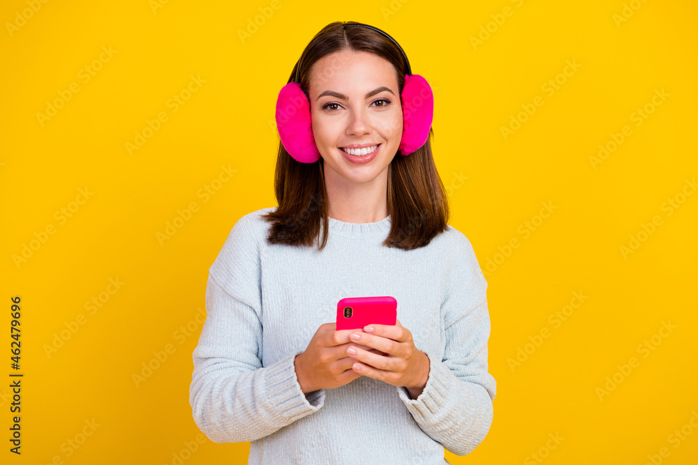 Photo of cheerful positive young woman wear earmuff hold phone repost comment isolated on yellow color background Photo of cheerful positive young woman wear earmuff hold phone repost comment isolated on yellow color background