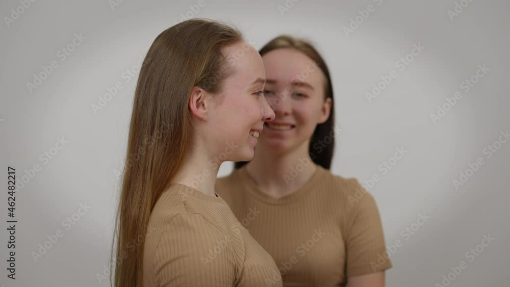 Laughing young woman posing with twin sister at background side view. Positive happy relaxed Caucasian identical women smiling having fun at grey background Laughing young woman posing with twin sister at background side view. Positive happy relaxed Caucasian identical women smiling having fun at grey background