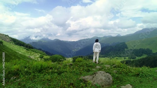 Aerial shot of woman hiker on top of mountain. Camera follows hipster young woman in white jacket running up on top of mountain summit.