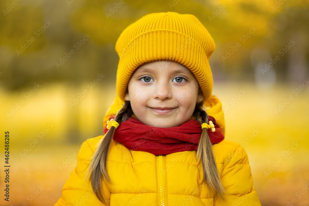Close up portrait of cute pretty little girl in yellow cap, jacket and red scarf, smiling at camera on beautiful golden autumn background outdoors in the park Close up portrait of cute pretty little girl in yellow cap, jacket and red scarf, smiling at camera on beautiful golden autumn background outdoors in the park