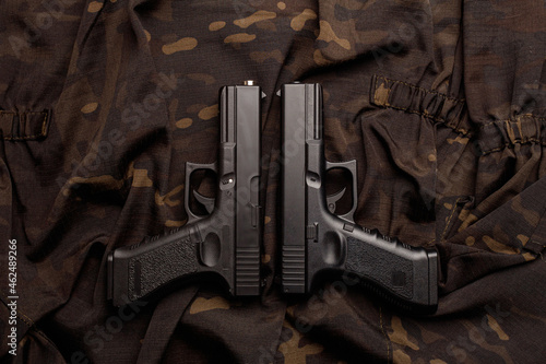 On a camouflage soldier's jacket there are two black pistols, the muzzles of which are pointed in one direction. A firearm that makes you think about self-defense. World Arms Day. Top view