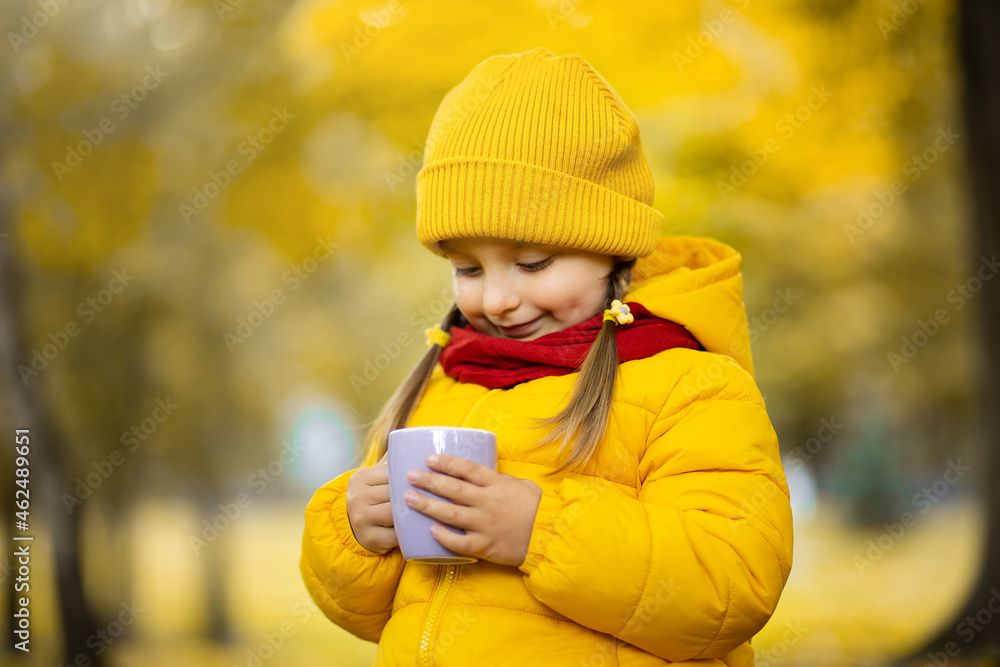 Warm fall photo. Close-up shot of a pretty cute little child girl wearing bright yellow coat and cap, holding a cup of hot chocolate or tea in autumn golden park Warm fall photo. Close-up shot of a pretty cute little child girl wearing bright yellow coat and cap, holding a cup of hot chocolate or tea in autumn golden park
