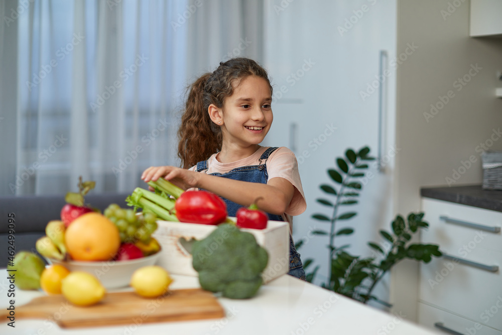 Adorable laughing and curly little girl sits at the table in the kitchen, near vegetables. Adorable laughing and curly little girl sits at the table in the kitchen, near vegetables.