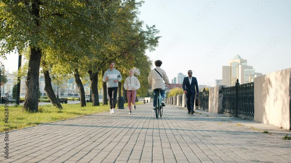 Diverse group of men and women are moving along urban street walking and by bike on summer day. Metropolitan landscape is in background. Diverse group of men and women are moving along urban street walking and by bike on summer day. Metropolitan landscape is in background.