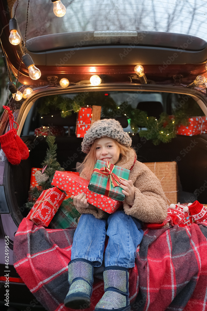  A seven-year-old girl wearing a red Santa Claus hat, sitting in a car decorated for the New Year, holds gifts prepared for friends under a Christmas tree