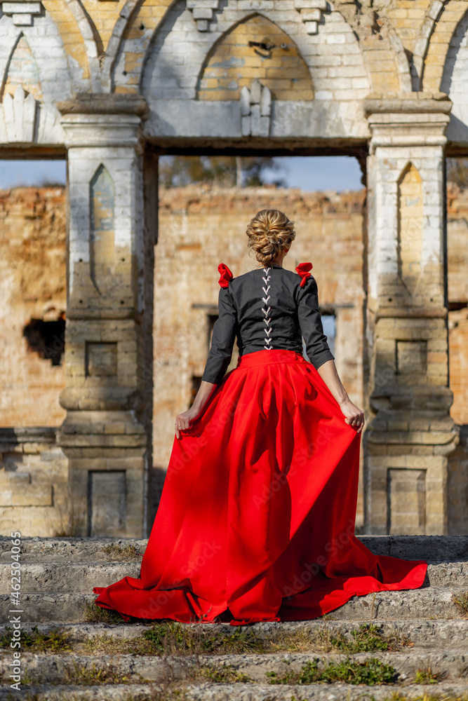 A woman in a 19th century dress in red and black walks to the ruins of an old castle manor in autumn