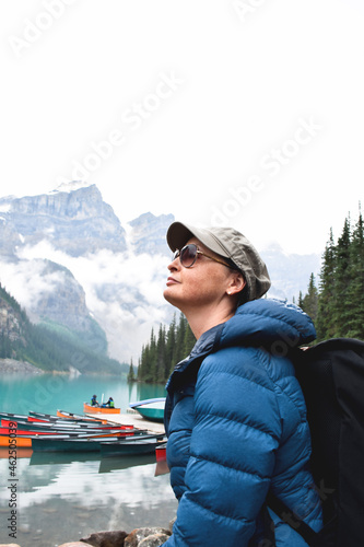 Closeup portrait of woman with backpack looking up with turquoise lake with colourful canoes and mountains behind her