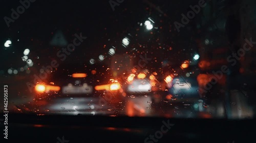 Wet car window with working brushes when driving through a traffic jam in the rain late at night.