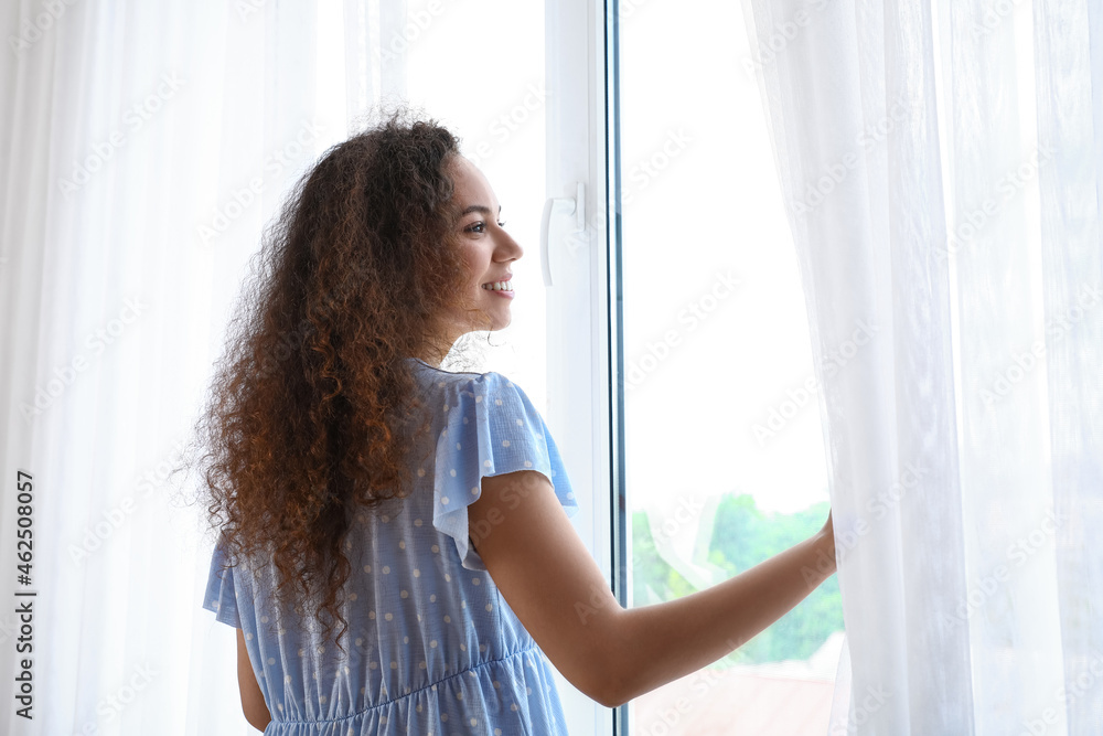 Young African-American woman opening light curtains at home in morning Young African-American woman opening light curtains at home in morning
