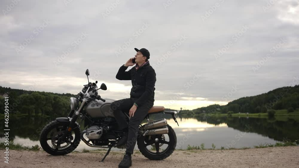 Motorbike driver talking on his phone sitting on a scrambler by lake in autumn. Guy sitting on a cafe racer by the lake. High quality 4k footage Motorbike driver talking on his phone sitting on a scrambler by lake in autumn. Guy sitting on a cafe racer by the lake. High quality 4k footage