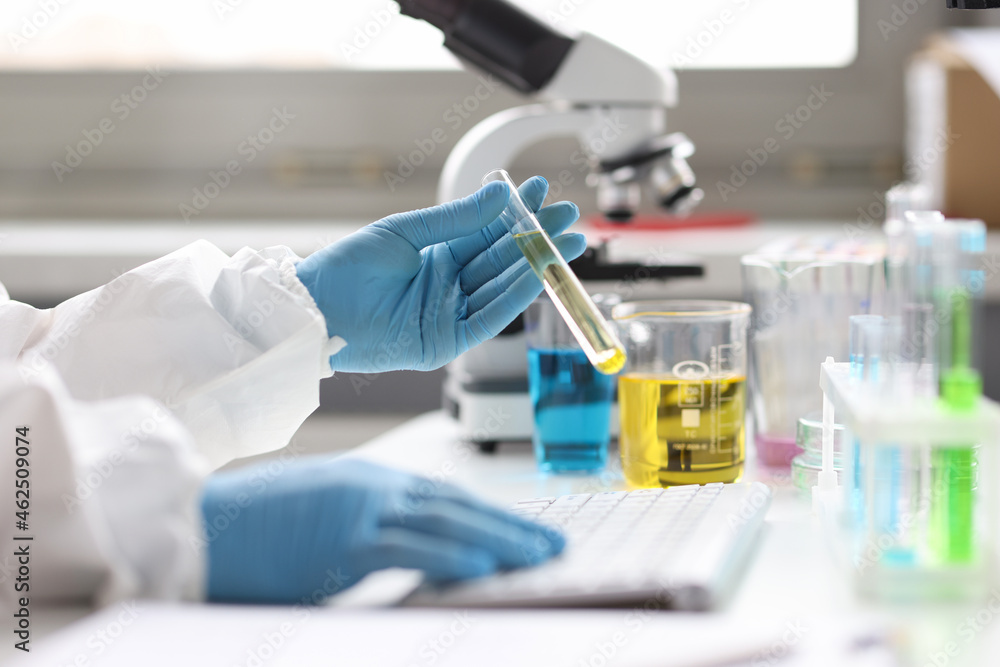 Gloved scientist holds test tube with transparent liquid and types on keyboard Gloved scientist holds test tube with transparent liquid and types on keyboard