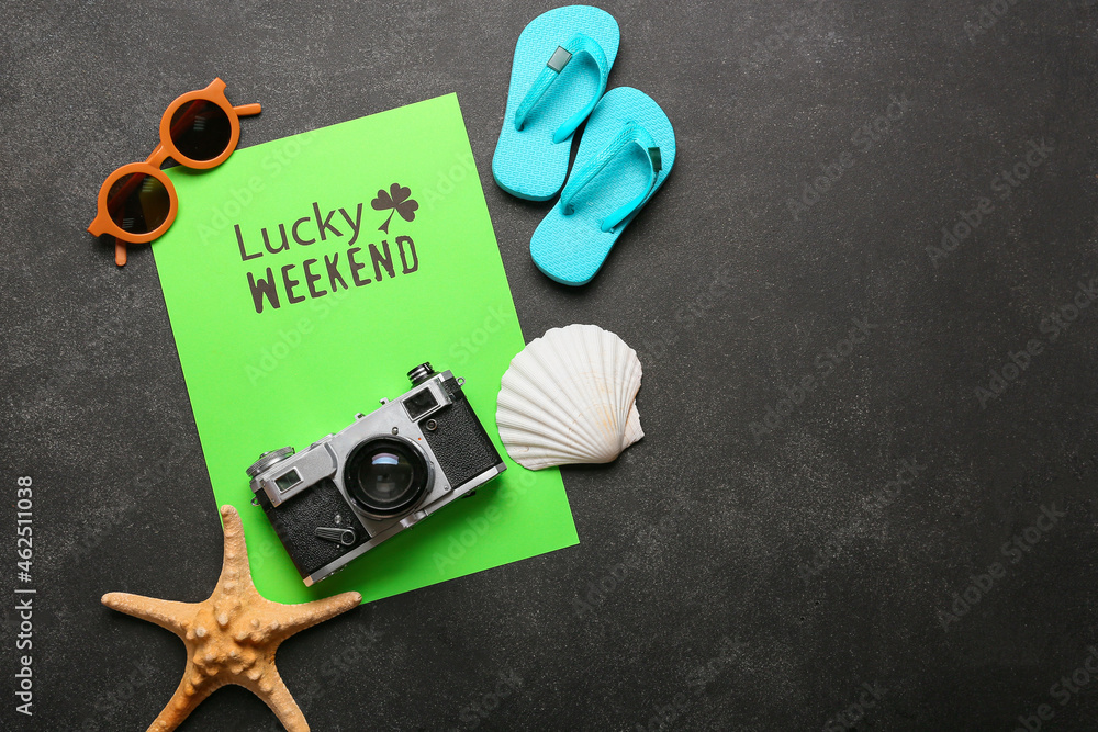Obraz premium Paper sheet with text LUCKY WEEKEND with photo camera, sunglasses and flip-flops on dark background. New year goals