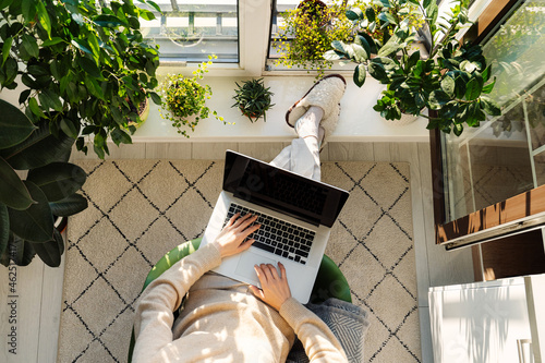 Freelancer woman working on laptop from home sitting at cozy balcony surrounded with green house plants. Businesswoman on remote occupation with computer at comfortable workplace near window. top view