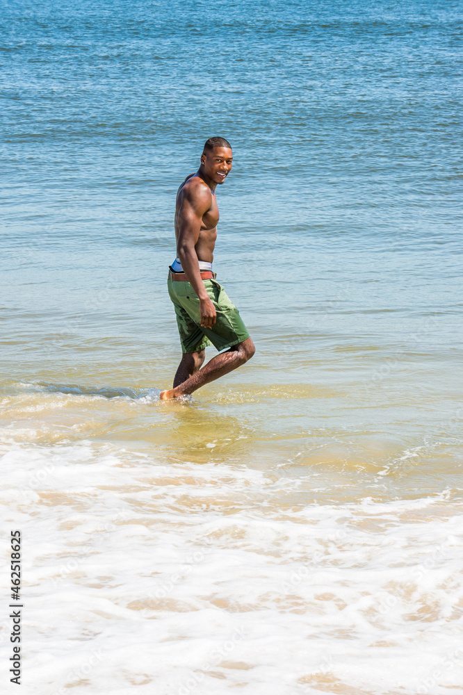 A well built young sexy black guy, half naked, is walking on water of ocean, turning back, smiling. .
