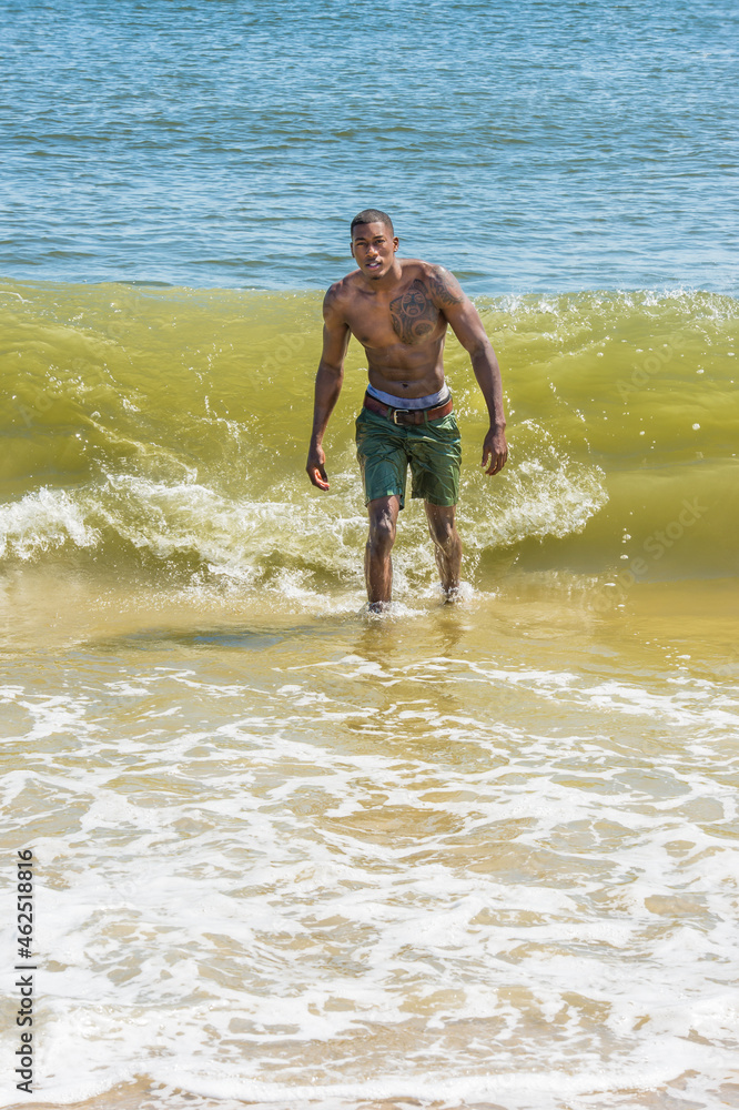 A well built young sexy black guy, half naked, is confidently walking out off ocean. Huge waves coming from back..