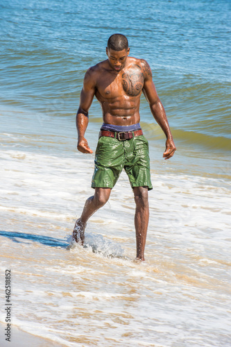A well built young sexy black guy, half naked and whole boy wet, is looking down and walking on the beach..