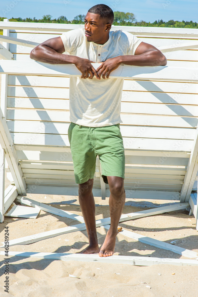 Bending over, both arms resting on a stick, a young handsome black guy is standing by a white wooden structure on the beach, looking around, relaxing..