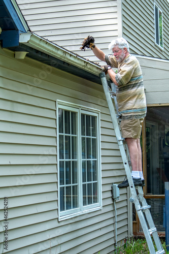 Man on  a ladder cleaning out gutters of his house © Susan