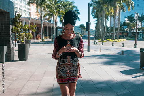 Beautiful African American woman wearing casula red and black dress and african turban standing up texting on the mobile cell phone and smiling. Palm trees and city buildings on the background. photo