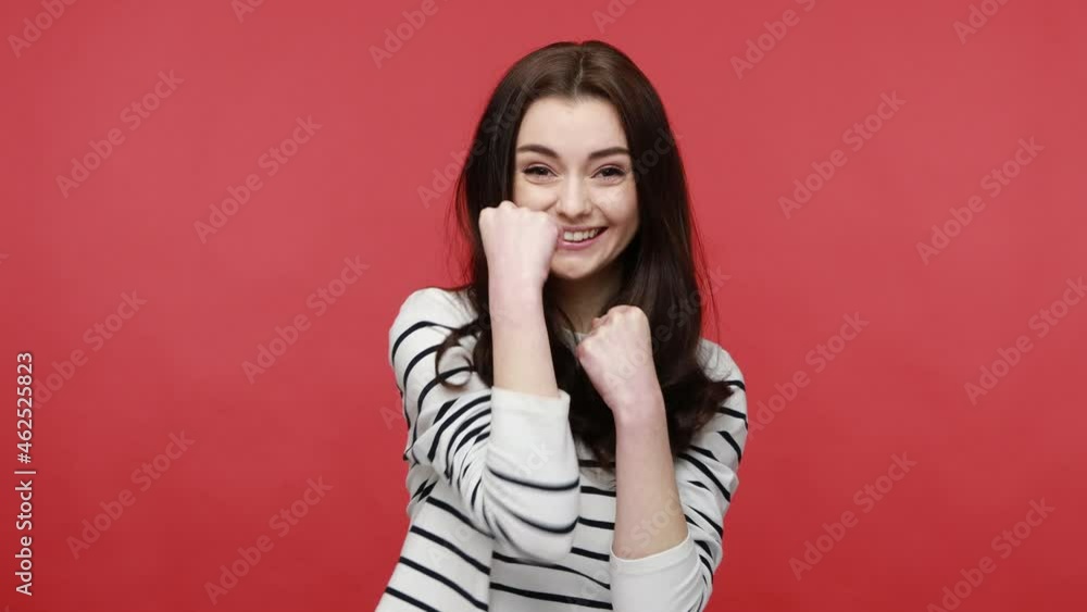 Woman standing with clenched fists, having fun, looking at camera with positive playful expression, wearing casual style long sleeve shirt. Indoor studio shot isolated on red background. Woman standing with clenched fists, having fun, looking at camera with positive playful expression, wearing casual style long sleeve shirt. Indoor studio shot isolated on red background.