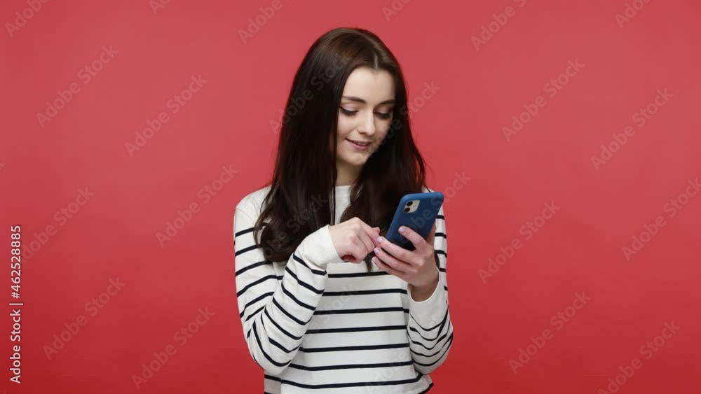Portrait of woman using cell phone, smiling while scrolling web, satisfied with mobile application, wearing casual style long sleeve shirt. Indoor studio shot isolated on red background.