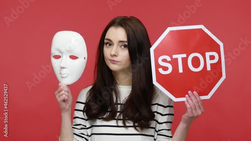 Portrait of attractive woman holding white mask and stop traffic sign to camera, nodding her head no, wearing casual style long sleeve shirt. Indoor studio shot isolated on red background. photo