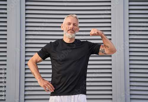 Confident middle aged man looking at camera, showing his bicep while posing outdoors over gray background