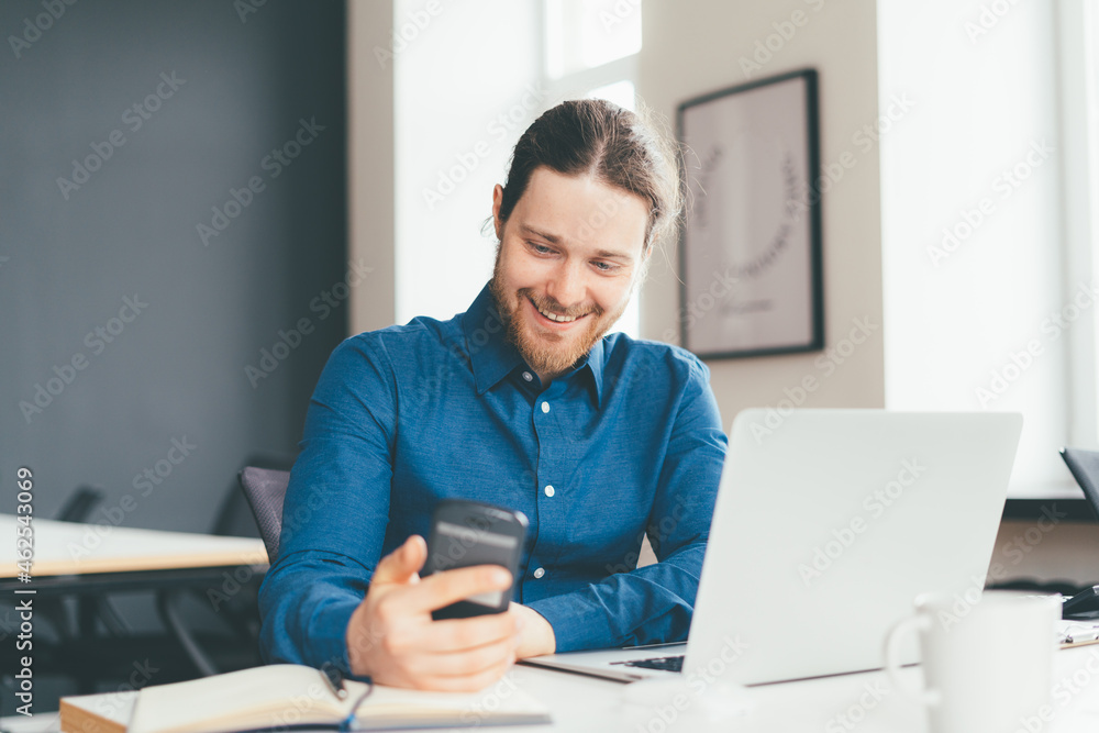 Smiling young male employee in casial clothes looking at smartphone while working on laptop in modern office centre. Happy manager using digital technologies at work Smiling young male employee in casial clothes looking at smartphone while working on laptop in modern office centre. Happy manager using digital technologies at work