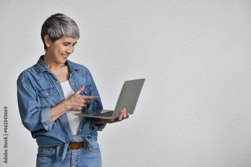 Portrait of middle aged Asian woman 50s wearing casual denim shirt white t-shirt holding laptop computer and pointing fingers isolated on white background Portrait of middle aged Asian woman 50s wearing casual denim shirt white t-shirt holding laptop computer and pointing fingers isolated on white background
