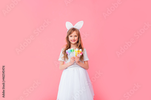 Cute blonde girl 8-9 years old in a white T-shirt and skirt with rabbit ears holds Easter eggs isolated on a pink background. Happy Easter, holidays, traditions.